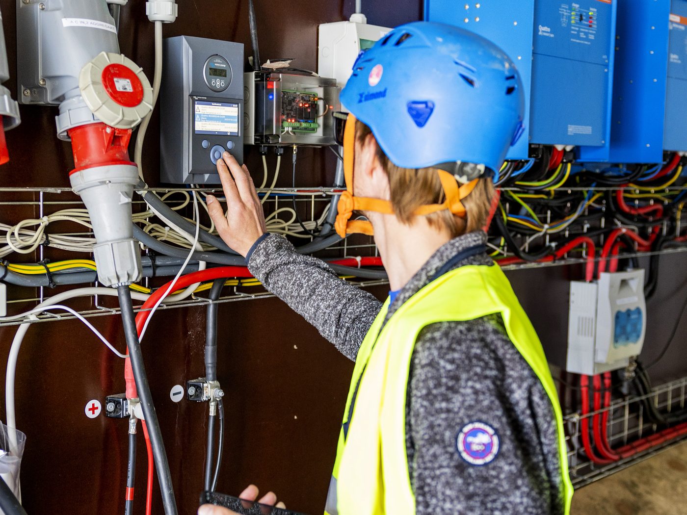 An engineer in a helmet and high-visibility vest working at a power cabinet full of inverters, breakers and heavy cabling